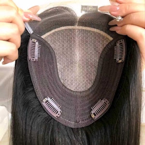 Pode incluir: Close-up de uma touca de cabelo preta com base de renda e clipes para fixação. O cabelo é longo e liso, com um brilho natural. Os clipes são de cor rosa claro.