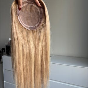 May include: A blonde hair topper with a circular base and multiple clips for attachment. The hair is straight and a light shade of blonde, with a natural-looking appearance. The base is a light brown color.