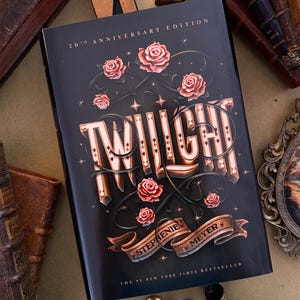 Pode incluir: A edição do 20º aniversário do livro Twilight de Stephenie Meyer apresenta uma capa preta com um título grande e estilizado em letras creme e douradas. O design inclui rosas cor-de-rosa, vinhas escuras e pequenas estrelas. O texto diz The #1 New York Times Bestseller.