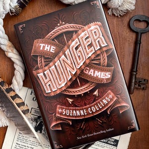 May include: A hardback book titled The Hunger Games by Suzanne Collins with a dark brown cover featuring a central circular emblem, decorative scrollwork, and arrows. The book is surrounded by a thick white rope, a dark metal key, and a patterned feather.