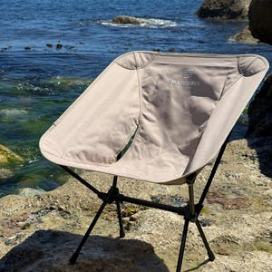 May include: A beige portable camping chair with a black metal frame. The fabric seat features the text MATCHBOX printed in white on the upper backrest. The chair has a curved, bucket-style design with reinforced corners and a collapsible structure.