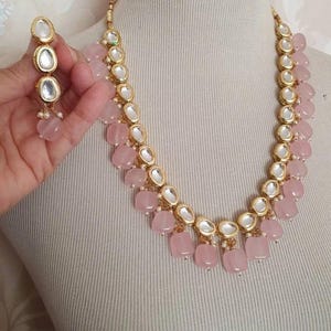 May include: A gold necklace featuring pink and white beads. The necklace has a layered design with larger pink beads at the bottom. The necklace is paired with matching earrings.