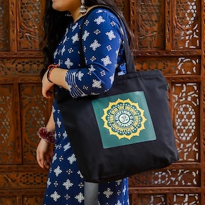 May include: Black tote bag with a square graphic featuring a yellow and green mandala design and the text "Into the Infinite, Everything is Connected, Divine Intelligence". The bag is carried by a person wearing a blue patterned dress.