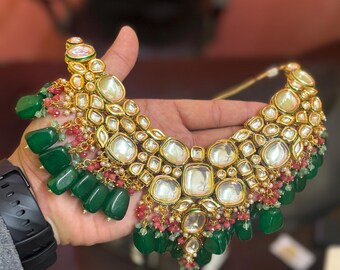 May include: A gold necklace with green and pink stones. The necklace is made of metal and has a traditional Indian design.