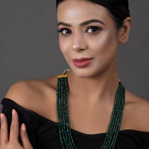 May include: A woman wearing a black off-the-shoulder top with a long, layered necklace made of dark green beads.