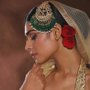May include: A woman wearing a gold and emerald green headpiece with a gold veil and two red roses. The headpiece has a crescent shape with dangling green beads and white pearls. The veil is a sheer gold fabric.