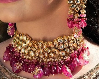 May include: A gold choker necklace featuring pink and white stones. The necklace has a layered design with multiple strands of beads and large, ornate pendants.