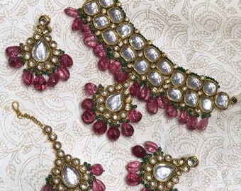 May include: A traditional Indian bridal jewellery set featuring a gold choker necklace, earrings, and a maang tikka. The necklace and earrings are adorned with large, round, clear stones and pink and green beads. The maang tikka is a small, delicate piece with a similar design.