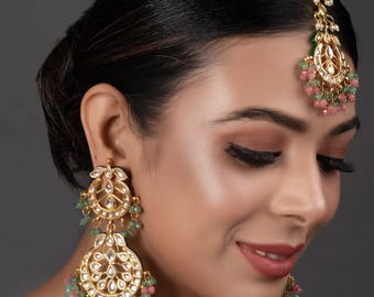 Peut inclure: Boucles d'oreilles et coiffe dorées avec des motifs complexes. Les bijoux présentent des pierres transparentes et des perles vertes et roses pendantes. Les boucles d'oreilles ont un design circulaire à plusieurs niveaux. La coiffe a un design similaire et se porte sur le front.