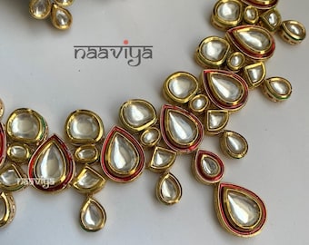 May include: A gold necklace with red and green accents features a teardrop-shaped design with multiple layers of clear stones. The necklace is made of metal and has a traditional Indian style.