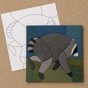 May include: A square fabric art piece featuring a stylized raccoon in shades of gray, black, and white, set against a blue sky and green ground. Beside the finished textile art is a white paper template showing the black outline of the raccoon.