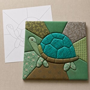 May include: A square fabric quilt block featuring a pieced turtle design in shades of teal, olive, and forest green. The turtle has a textured teal shell and patterned fabric body segments. A white paper pattern template with black outlines is included.