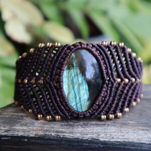 May include: A wide, dark brown macrame bracelet with a large, oval labradorite stone at its centre. The stone has a mix of blue, green, and grey hues. Small gold-coloured beads accent the macrame design.