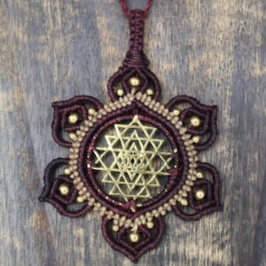 May include: A handcrafted macrame pendant necklace featuring a geometric gold-colored design at the center. The pendant is a deep burgundy color with gold-colored accents and a matching cord.