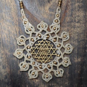 May include: Cream-colored macrame necklace with a gold-colored pendant featuring a geometric design. The necklace has a circular design with small gold beads and a cream-colored cord. The pendant is a detailed geometric pattern.