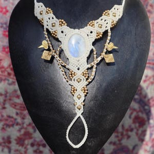 May include: A handmade macrame necklace with a large oval moonstone pendant. The off-white necklace features gold-colored beads and decorative gold charms. The necklace is displayed on a black mannequin.
