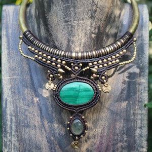 May include: A handcrafted necklace featuring a large oval malachite stone pendant. The necklace has a bronze-colored collar and is adorned with intricate macrame knots, small gold beads, and decorative gold accents. The lower pendant is a smaller, oval green stone.
