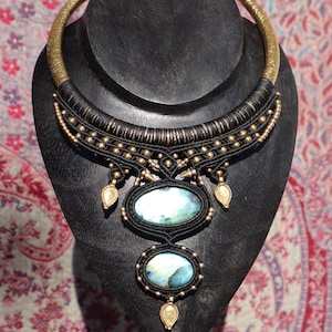 May include: A handcrafted statement necklace featuring a wide, gold-toned collar and intricate black macrame detailing. Two oval, iridescent stones are suspended from the macrame, accented with gold-colored beads and teardrop pendants. The necklace is displayed on a black jewelry stand.