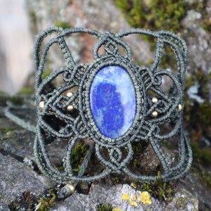 Könnte beinhalten: Ein dunkelgrünes Makramee-Armband mit einem großen ovalen Lapislazuli-Edelstein in der Mitte. Das Armband ist mit kleinen goldenen Perlen verziert.
