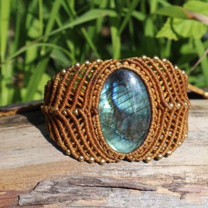 May include: A macrame bracelet featuring a large, oval-shaped labradorite gemstone with blue and green iridescence at its center. The band is crafted from brown cord in a complex knotted pattern, accented with small, round, metallic beads throughout.