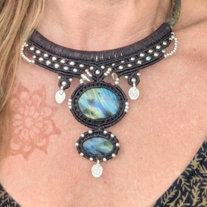 May include: A black macrame necklace with two oval labradorite stones. The necklace has silver accents and a curved design. The labradorite stones display iridescent blue and green hues. The necklace is worn on a person.