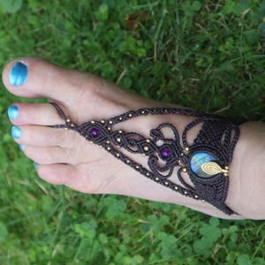 Può includere: Sandali a piedi nudi in macramè marrone scuro con perline dorate e pietre preziose viola e blu. I sandali sono indossati su un piede con smalto blu, su uno sfondo di erba verde.