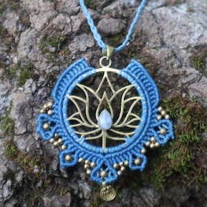 May include: A blue macrame necklace with a gold-colored lotus flower pendant. The pendant features a small, light-colored stone at its center and is adorned with gold-colored beads. The necklace is displayed against a textured, natural background.