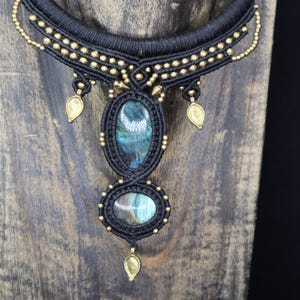 May include: A handcrafted macrame necklace featuring a black cord design with gold-colored accents. The necklace showcases two oval-shaped, iridescent labradorite stones. The necklace has a brass-colored collar.