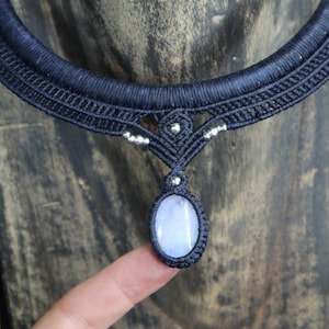 May include: A dark blue macrame necklace with a central pendant. The pendant holds a light blue oval stone. The necklace is adorned with silver-coloured beads. The background is a wooden surface. The necklace is handmade.
