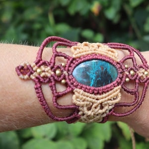 May include: A handmade macrame bracelet featuring a central oval stone with blue and green hues. The bracelet is crafted with burgundy and tan colored cords, accented with small gold-colored beads. The design is intricate and bohemian in style.