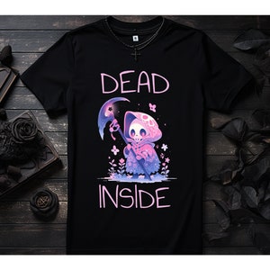 Puede incluir: Camiseta negra con un gr&aacute;fico rosa de un esqueleto de dibujos animados que lleva una capa con capucha y sostiene una guada&ntilde;a. El texto "DEAD INSIDE" est&aacute; impreso en rosa debajo del gr&aacute;fico.