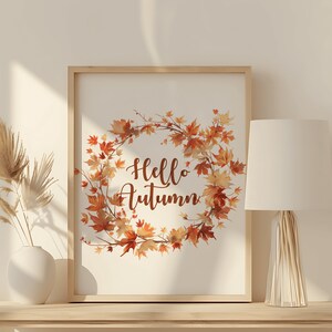 Puede incluir: Una impresi&oacute;n enmarcada con una ilustraci&oacute;n de acuarela de una corona de hojas de oto&ntilde;o y el texto "Hello Autumn". La impresi&oacute;n se muestra en un estante de madera clara con una l&aacute;mpara blanca y un jarr&oacute;n con hierba de la pampa seca.