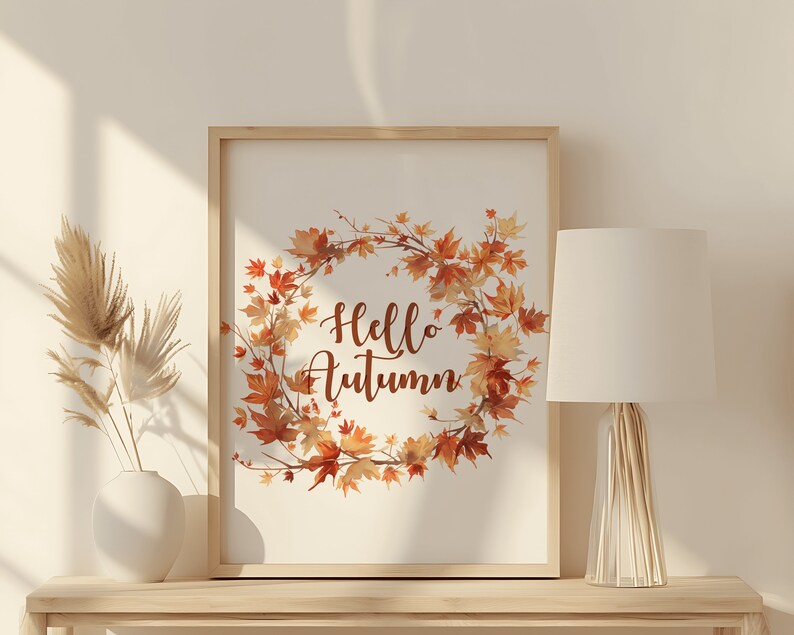 Puede incluir: Una impresi&oacute;n enmarcada con una ilustraci&oacute;n de acuarela de una corona de hojas de oto&ntilde;o y el texto "Hello Autumn". La impresi&oacute;n se muestra en un estante de madera clara con una l&aacute;mpara blanca y un jarr&oacute;n con hierba de la pampa seca.