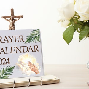May include: A white prayer calendar with the words "PRAYER CALENDAR 2024" printed on the front. The calendar features a watercolor illustration of a hand reaching up towards a bright light in the clouds. A small golden cross with a figure of Jesus Christ is on top of the calendar.