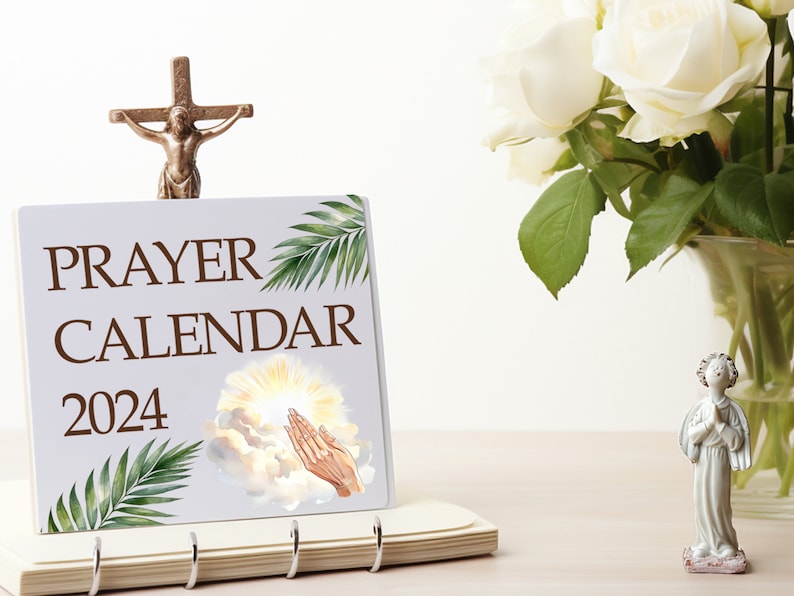 May include: A white prayer calendar with the words "PRAYER CALENDAR 2024" printed on the front. The calendar features a watercolor illustration of a hand reaching up towards a bright light in the clouds. A small golden cross with a figure of Jesus Christ is on top of the calendar.