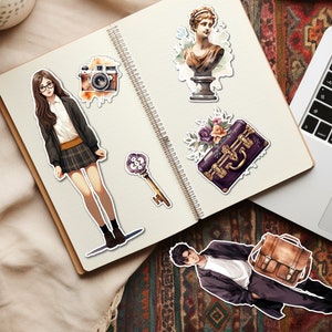 Pode incluir: Um caderno com v&aacute;rios adesivos, incluindo uma mulher de &oacute;culos, uma c&acirc;mera, uma chave, um busto de mulher, uma mala com flores e um homem de terno. O caderno est&aacute; em um tapete estampado.