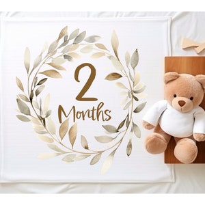 May include: White baby blanket featuring a brown watercolour wreath with the text "2 Months". A brown teddy bear is sitting on a wooden surface beside the blanket.