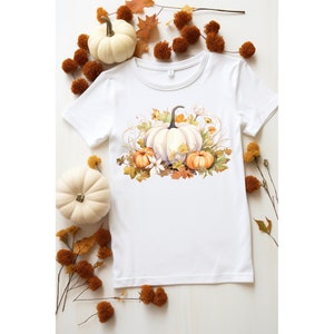 以下が含まれることがあります： 白のTシャツ。水彩画のデザインで、白、オレンジ、黄色のカボチャ、葉、花が描かれています。シャツの周りには小さな白いカボチャと秋の葉があり、季節感のあるテーマになっています。