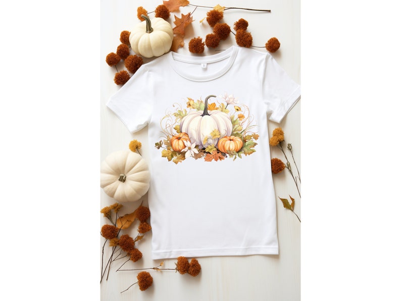 以下が含まれることがあります： 白のTシャツ。水彩画のデザインで、白、オレンジ、黄色のカボチャ、葉、花が描かれています。シャツの周りには小さな白いカボチャと秋の葉があり、季節感のあるテーマになっています。