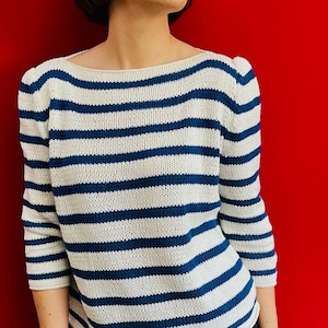 May include: A woman with short, dark hair is wearing a white and blue striped sweater with a boat neck. She is standing in front of a red background.