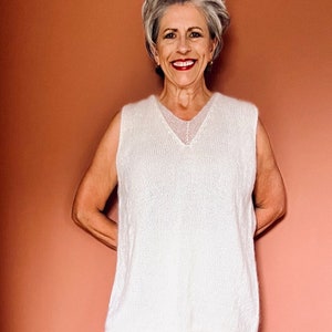 Könnte beinhalten: Eine Frau in einem cremefarbenen Outfit. Das Outfit besteht aus einem ärmellosen V-Ausschnitt-Pullover und einer Hose mit weitem Bein. Die Frau trägt auch weiße Sandalen. Der Hintergrund ist einfarbig, gedämpftes Orange.