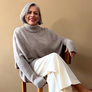 May include: A woman wearing a light grey turtleneck sweater and white trousers. She is sitting on a wooden stool with her legs crossed.