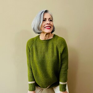 Könnte beinhalten: Eine Frau trägt einen grünen Strickpullover mit weißen Ärmelbündchen und eine weiße Hose.