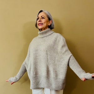 Puede incluir: Una mujer con un suéter gris claro de cuello alto de corte holgado y pantalones blancos.