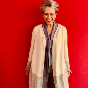 Peut inclure: Une femme portant un long cardigan crème avec une bande grise en bas. Elle porte également une écharpe grise et un pantalon gris.