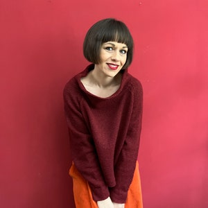 May include: A woman wearing a maroon sweater and orange corduroy trousers stands in front of a red wall.