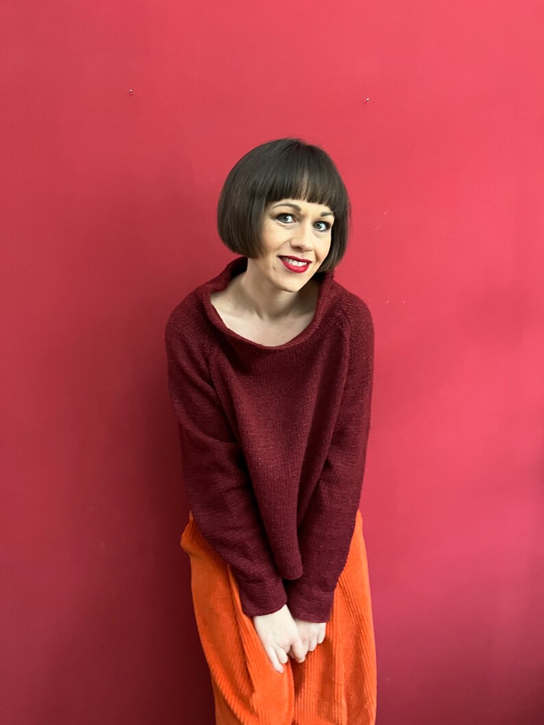 May include: A woman wearing a maroon sweater and orange corduroy trousers stands in front of a red wall.