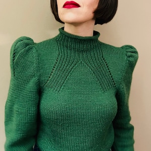 May include: A green knitted sweater with a high neck and puffed sleeves. The sweater has a delicate stitch pattern and is worn over a blue and green plaid skirt.
