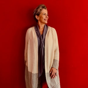 Könnte beinhalten: Eine Frau trägt einen langen, cremefarbenen Cardigan mit einem grauen Streifen am Saum. Sie trägt darunter ein graues Oberteil und hat kurze, graue Haare.