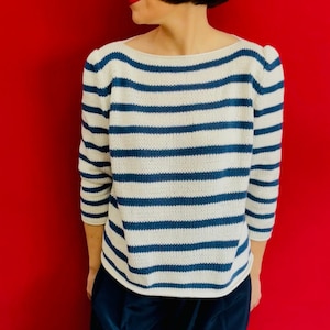 May include: A woman wearing a white and blue striped sweater with a navy blue pair of pants. She is standing in front of a red background.